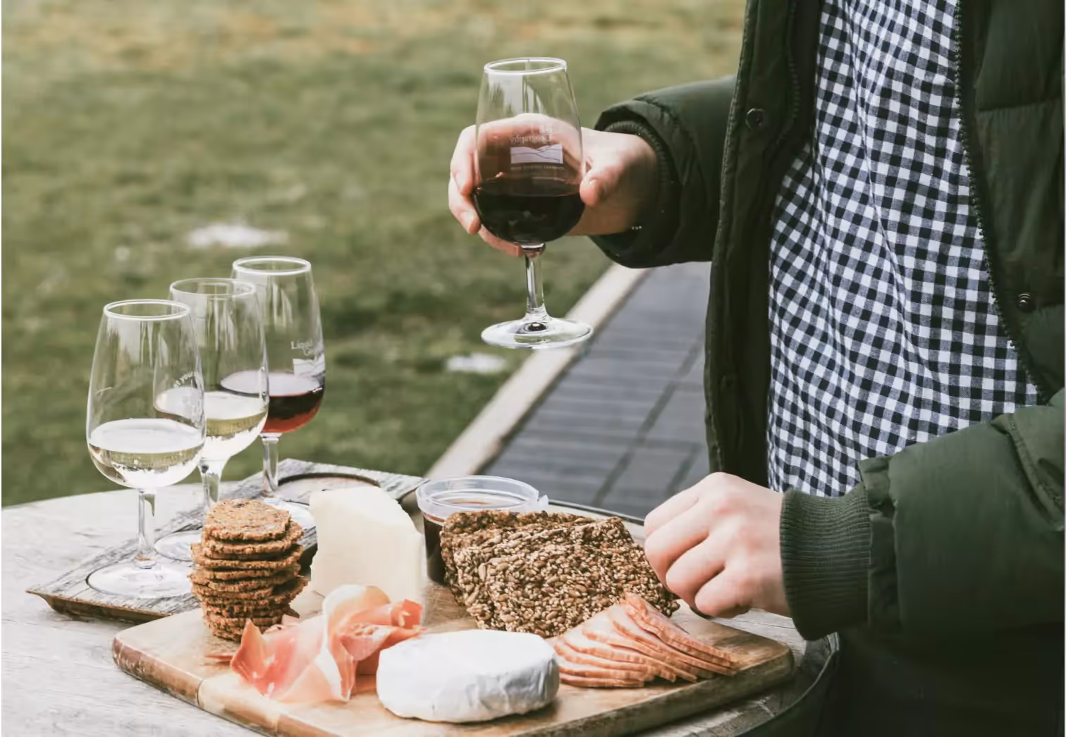 a person holding a glass of red wine over a table with glasses of red and white wine and a grazing platter of cheese meats and crackers