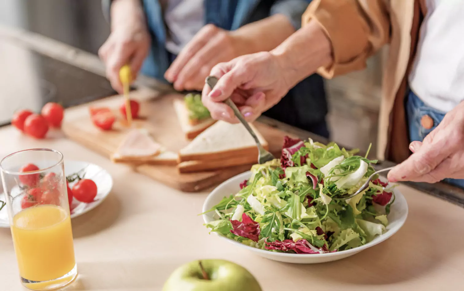 two people in the kitchen one of them making a sald, another making sandwiches