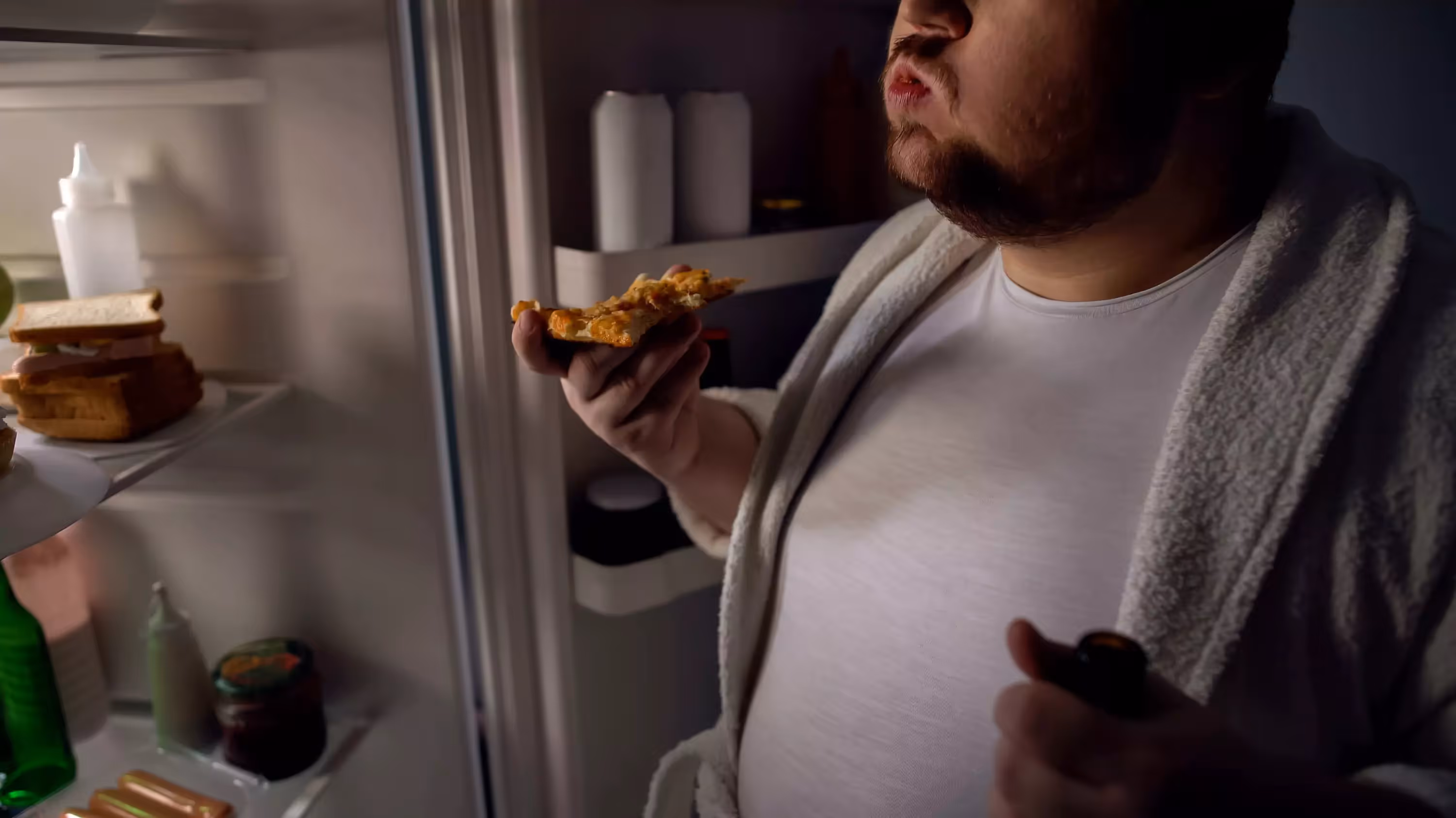 a person near an open fridge holding a slice of pizza and a bottle of beer 