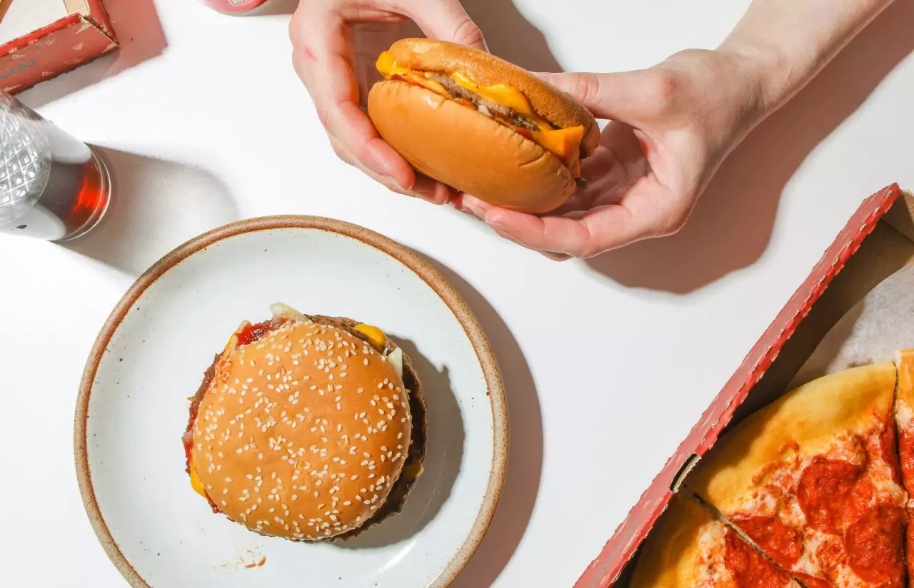 A person holding a cheeseburger next to a plate with a cheeseburger and a box of pepperoni pizza