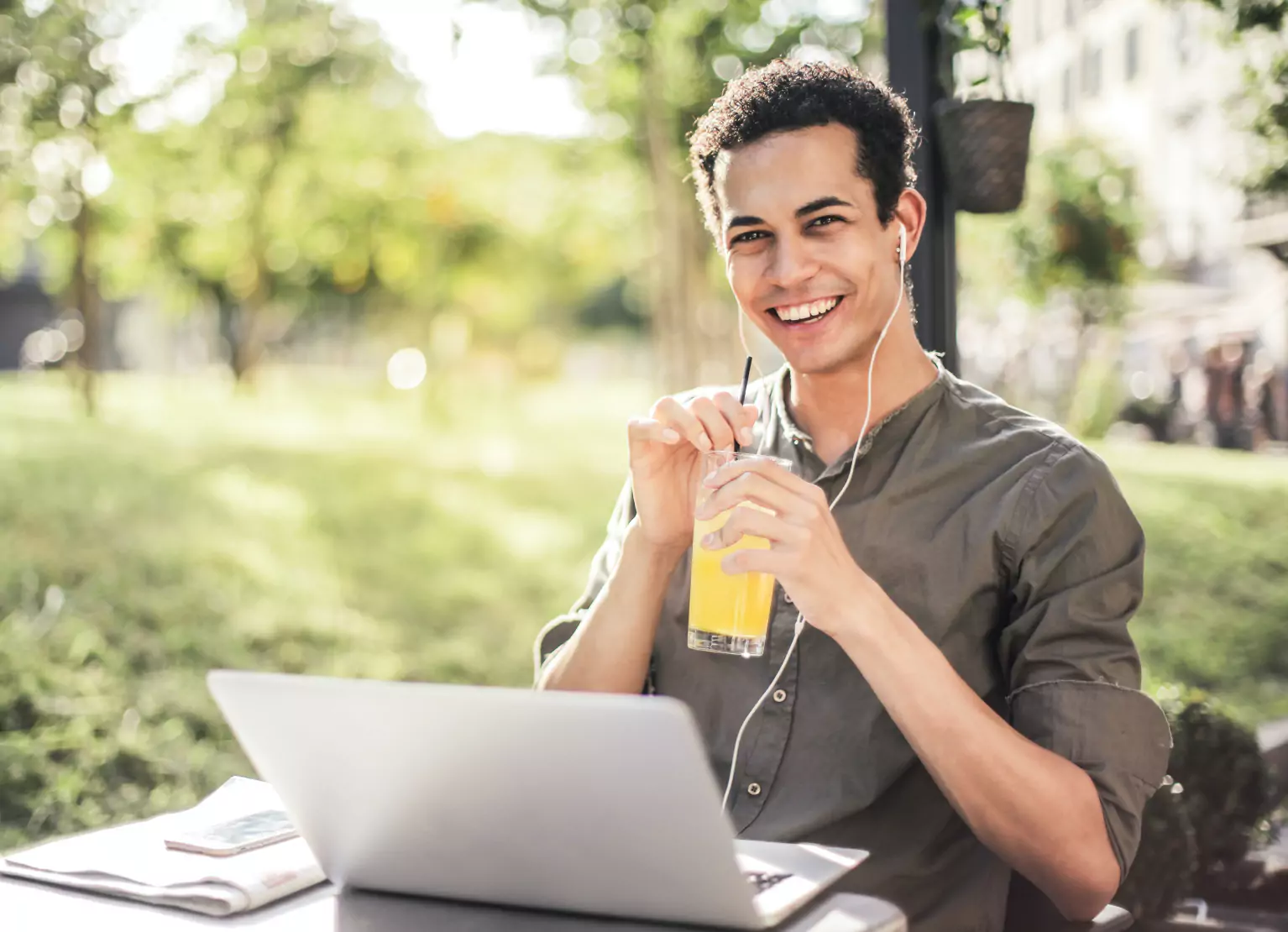 A person sitting outdoors with a laptop, wearing earphones and drinking orange juice